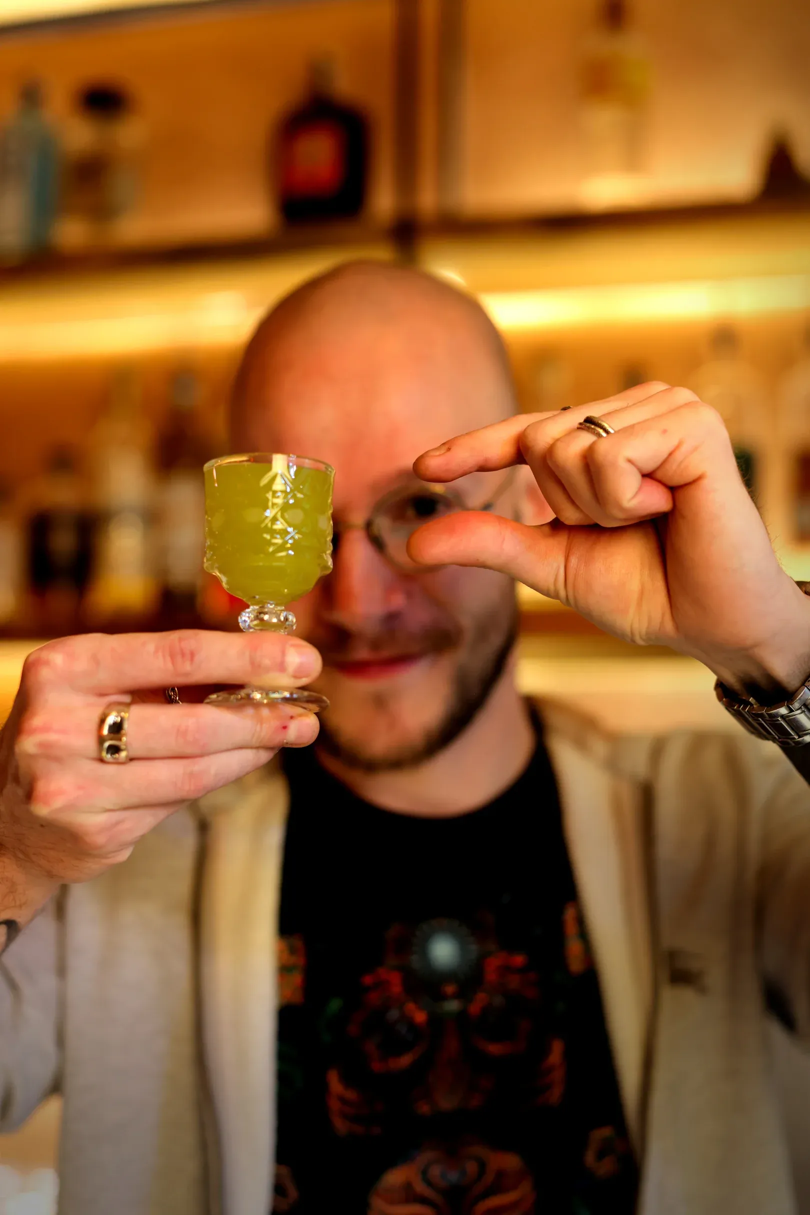 The bartender sizing up a green cocktail in a stemmed glass, one eye peeking around it