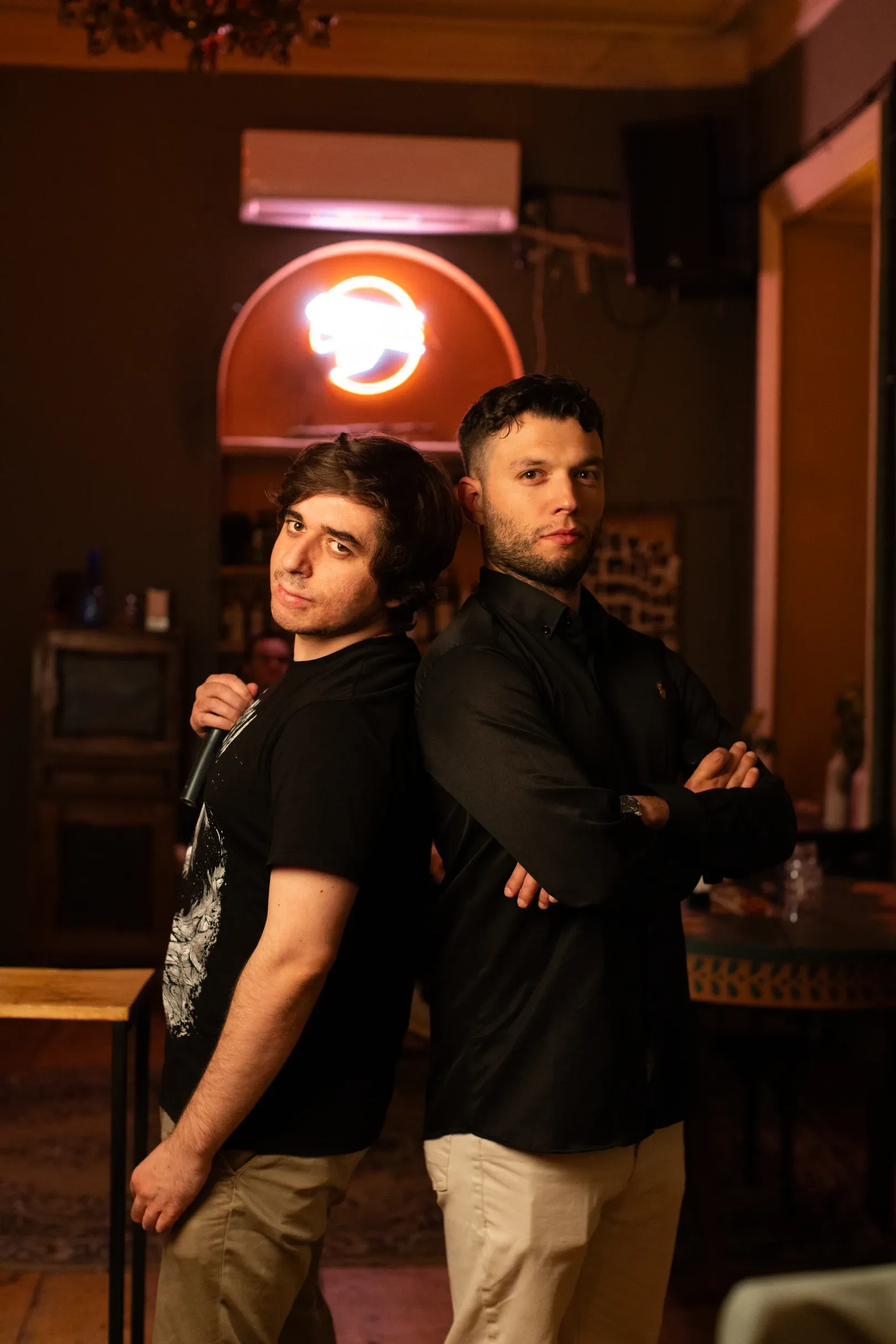 The two karaoke hosts posing back to back under the neon sign, microphone in hand