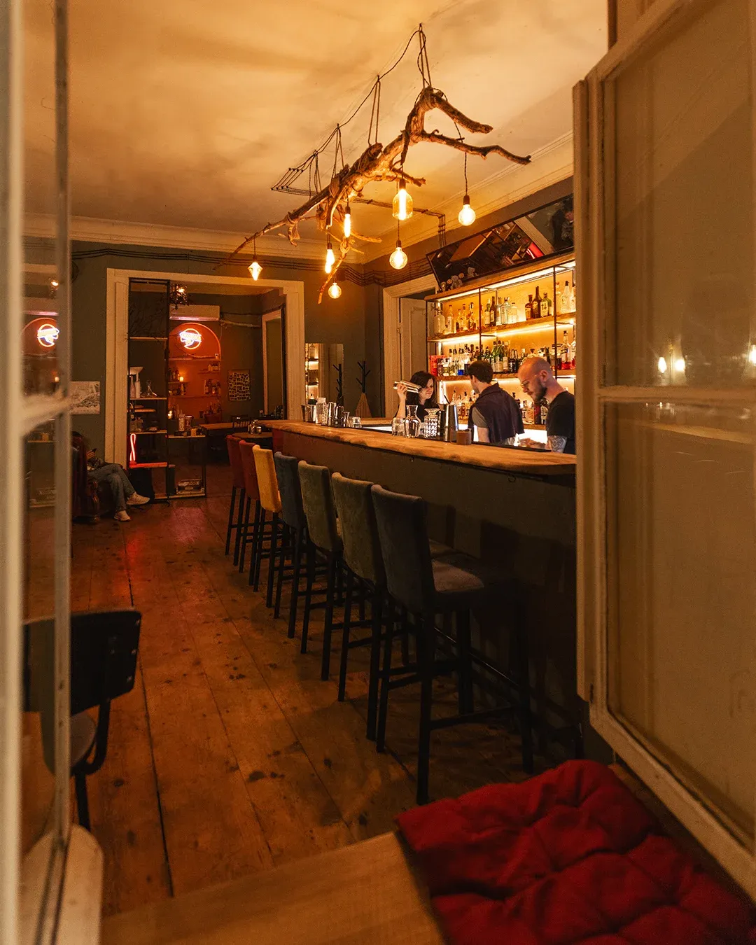 Looking down the bar room through an open window: the branch chandelier, velvet stools, and the lit back-bar