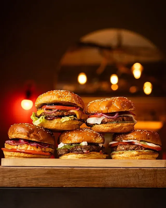 Five sesame-bun burgers stacked on a wooden board, bar lights glowing behind