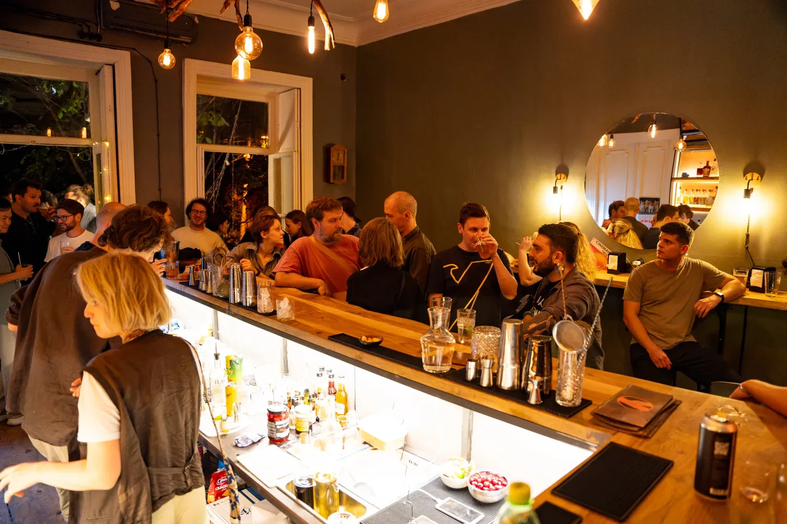 The room at full meetup: guests in conversation across the bar counter under warm bulbs
