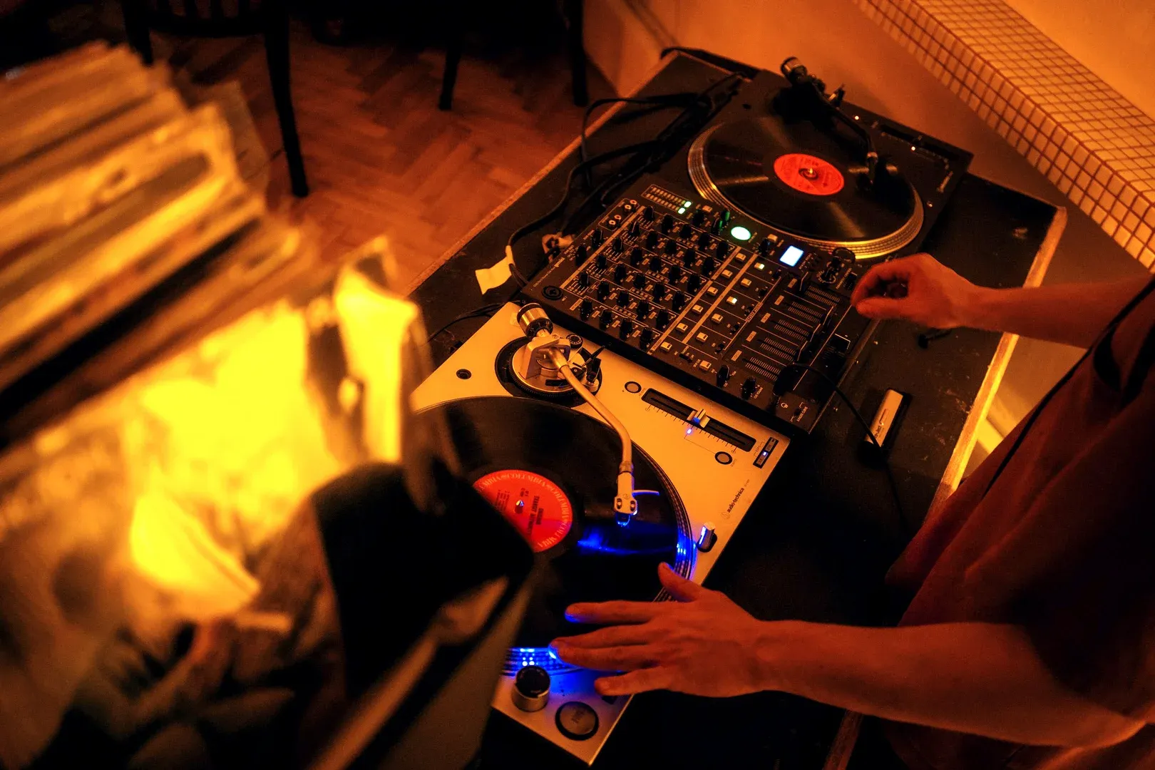 A DJ's hands on the decks: two turntables, a mixer, and a crate of records at their side