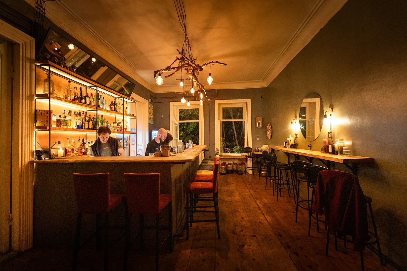 The bar counter at Crossroads: staff at work, lit back-bar shelves, red stools down the room